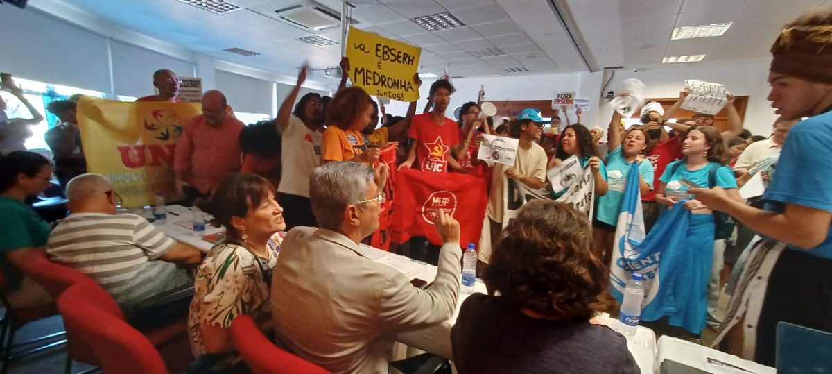 Foto feita de trás da mesa do Consuni, mostrando conselheiros de costas e, à frente, manifestantes contrários à votação