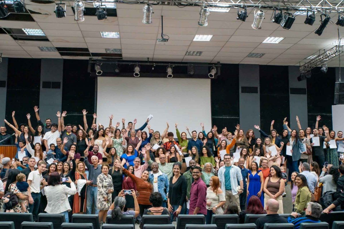 Fotografia de dezenas de pessoas levantando as mãos em sinal de comemoração em um auditório.