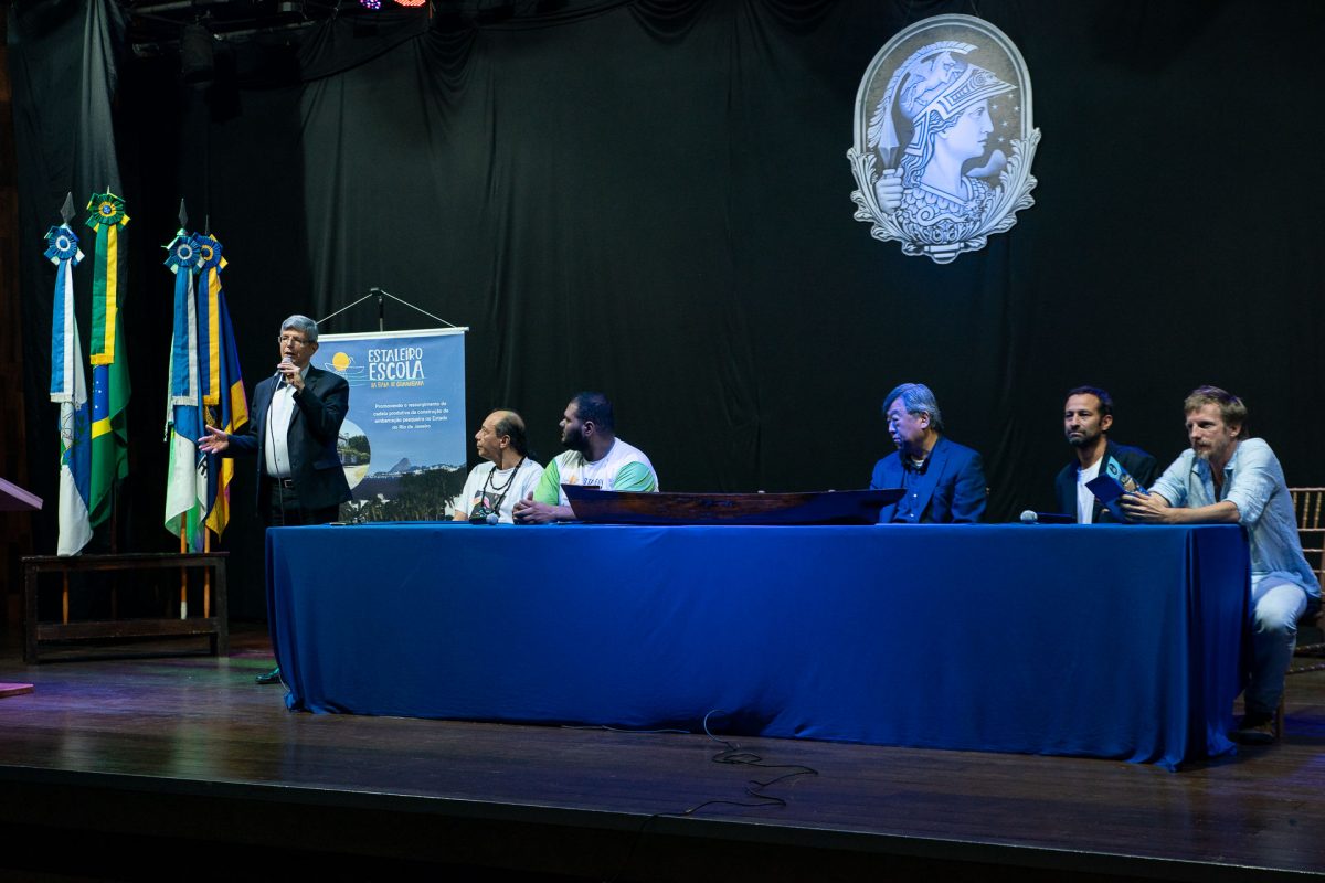 Fotografia de um palco. No fundo preto está a minerva da UFRJ e um banner do evento I Seminário Estadual de Fortalecimento da Cadeia Produtiva das Embarcações de Pesca e Fomento à Construção Naval Artesanal. Do lado direito está Roberto Medronho, falando ao microfone. Compondo a mesa de abertura do evento estão quatro homens: Walter Suemitsu, Luan Guimarães, Maurício Oliveira e Sérgio Ricardo.