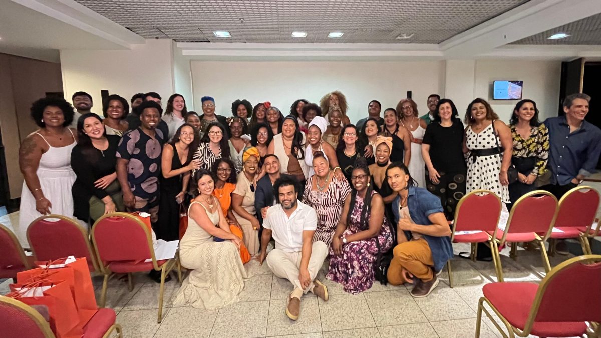 Fotografia que mostra um grupo de pessoas reunidas na formatura do curso de extensão Pretonomia. À frente do grupo, sentado no chão, está o professor Breno Cruz, idealizador do curso. Ele é um homem de pele negra, tem cabelos e barba escuros, veste camisa branca e calça bege.