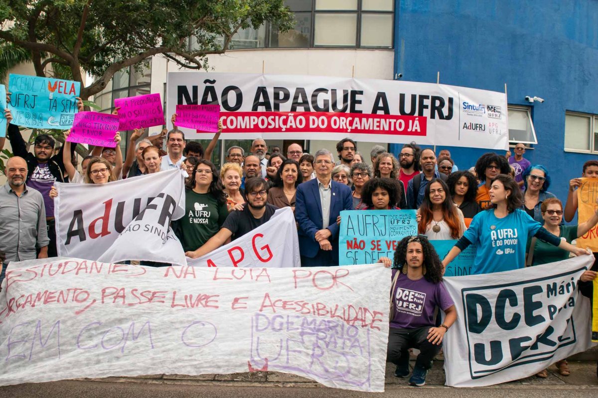 Fotografia mostra grupo de pessoas após sessão do Conselho Universitário (Consuni). Entre elas, a vice-reitora da UFRJ, Cássia Turci; o reitor da Universidade, Roberto Medronho; além de professores, estudantes e servidores técnico-administrativos. Eles seguram cartazes e faixas. Em uma das faixas, está escrito: “Não apague a UFRJ. Recomposição do orçamento já”.