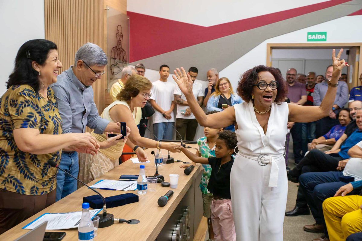 Numa fotografia realizada durante cerimônia em homenagem aos servidores com 50 anos de serviços públicos prestados à UFRJ, uma servidora comemora com os braços erguidos no momento em que se aproxima da mesa formada pelo reitor, Roberto Medronho, pela vice-reitora, Cássia Turci, e pela pró-reitora de pessoal, Neuza Luzia Pinto.