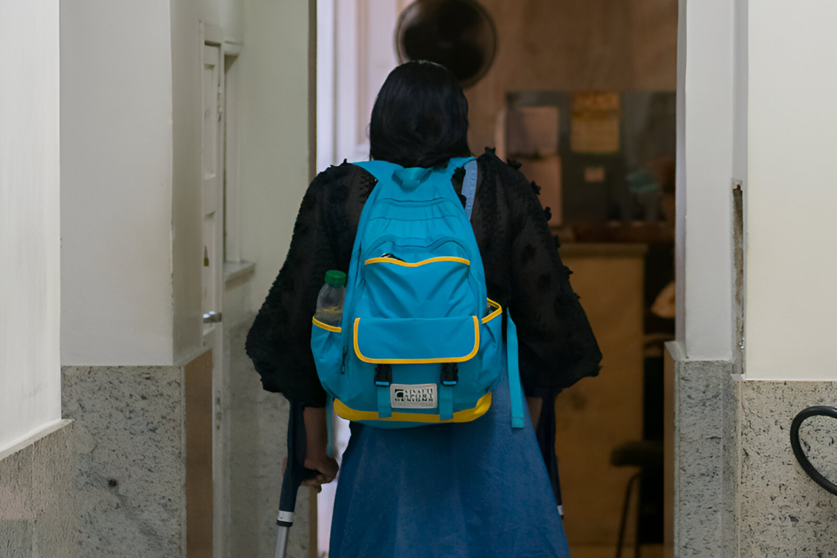 Fotografia de uma mulher de costas caminhando com o auxílio de muletas. Ela usa saia jeans, blusa preta e carrega uma mochila azul nas costas.