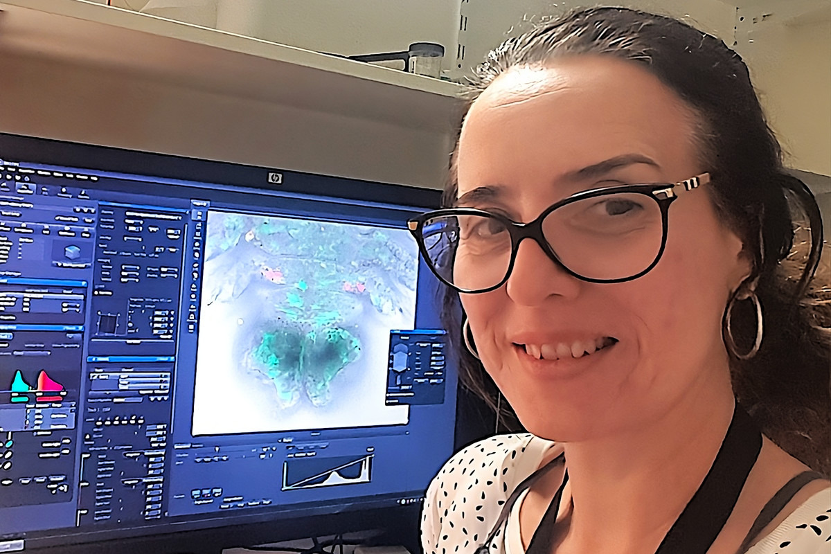 Fotografia que mostra, em primeiro plano, a professora Christine Ruta sorrindo discretamente. Ela é uma mulher branca, com cabelos escuros na altura dos ombros, usa óculos e brincos de argola.