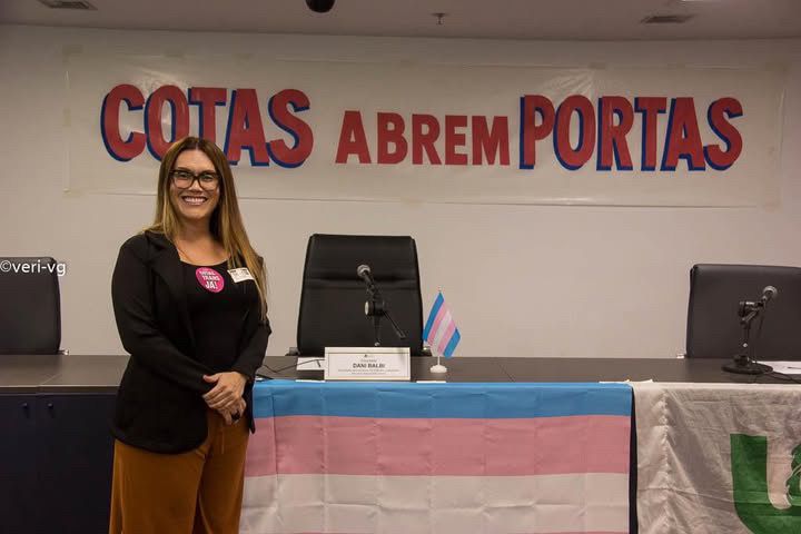 Uma mulher travesti está posta diante de uma mesa onde há a bandeira trans estendida. Atrás há uma placa onde se lê "COTAS ABREM PORTAS".