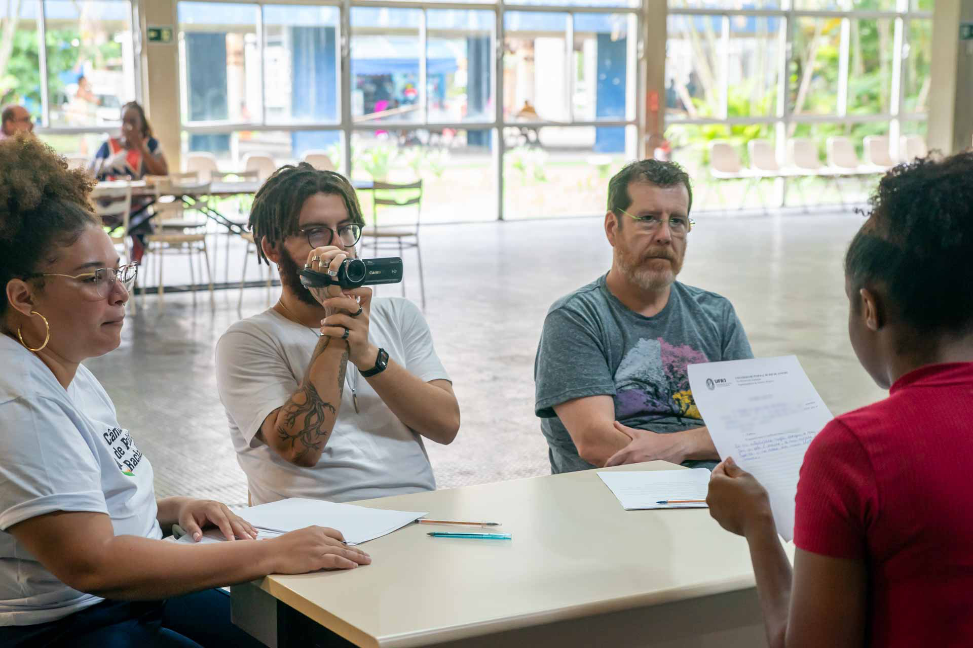 Fotografia de quatro participantes de uma banca de heteroidentificação. Eles estão sentados ao redor de uma mesa: duas mulheres e dois homens. A candidata lê o termo de autodeclaração étnico-racial enquanto é filmada por um dos componentes.