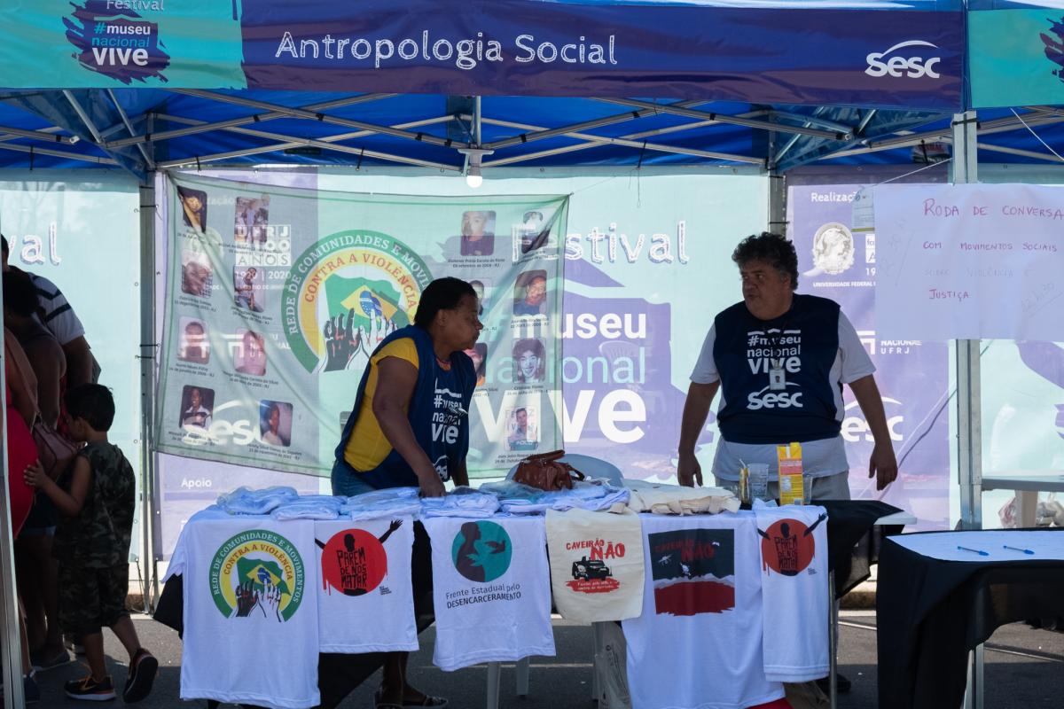 Estande do Festival Museu Nacional Vive em azul com os dizeres "Antropologia Social" e duas pessoas no centro organizando camisetas na bancada