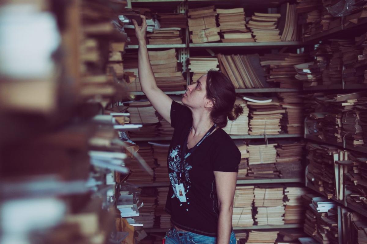 Aluna da UFRJ cercada por livros antigos em uma biblioteca. Ela se estica para retirar um livro na parte esquerda da imagem.