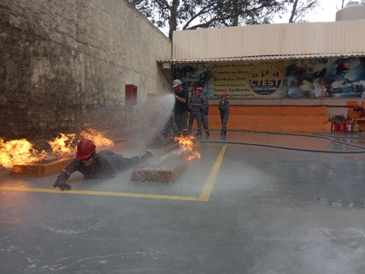 Mulher com roupa de combate à incêndio rasteja entre chamas enquanto outras pessoas jogam água com mangueira de incêndio durante simulação