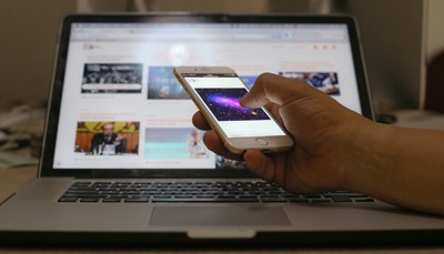 Imagem em primeiro plano de uma mão segurando um celular, na frente de um laptop, cujo teclado está embaixo da mão e cuja tela está atrás, em segundo plano e desfocado. A imagem representa o isolamento social provocado pelas redes sociais.