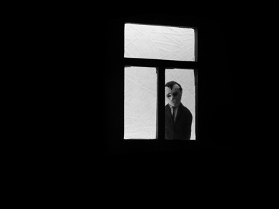 Ilustração em preto e branco de uma pessoa de terno e gravata pretos, trancada em seu quarto e olhando pela janela.