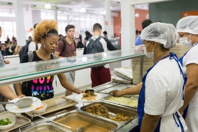 Na fila do Restaurante Universitário, trabalhadoras da alimentação servem estudantes. No prato, carne, macarrão e salada.