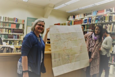 No interior de uma biblioteca, rodeadas por estantes de livros, duas pessoas carregam um grande mapa.