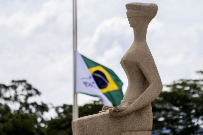Em primeiro plano, a imagem mostra a escultura de uma mulher com os olhos vendados e uma espada nas mãos, representando a justiça brasileira. Em segundo plano, a bandeira do Brasil aparece em meia-haste.