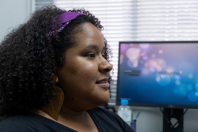 Uma jovem mulher negra está posicionada de perfil, conversando. Ao fundo, uma tela de computador exibe imagens aleatórias