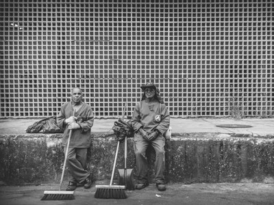 Foto em preto e branco de dois trabalhadores da limpeza, uniformizados e com instrumentos de trabalho nas mãos, em horário de descanso